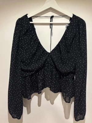 Svart blus med vita prickar och v-ringning - Snygg svart blus med vita prickar, djup v-ringning och knytband i nacken. Blusen har lång ärm och volang nedtill, tillverkad i ett tunt, lätt material som ger ett luftigt intryck. 