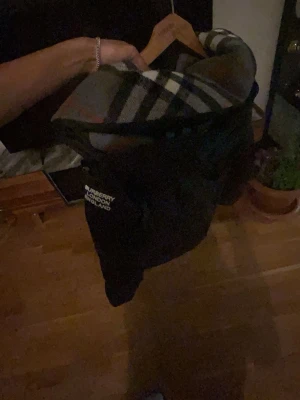 Svart pufferjacka från Burberry - Säljer en svart pufferjacka från Burberry med klassiskt rutigt foder i huvan. Jackan har dragkedja och knappar framtill samt två sidofickor. Perfekt för kalla vinterdagar och riktigt snygg med den ikoniska Burberry-looken.