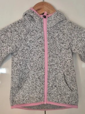 Gråmelerad hoodie med rosa dragkedja - Mysig gråmelerad hoodie med huva och rosa dragkedja. Tröjan har långa ärmar, fickor framtill och rosa kantband vid ärmslut och nederkant. Perfekt för kyliga dagar och enkel att matcha med andra plagg.i storlek 92 