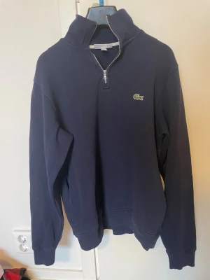 Marinblå halvzip från Lacoste - Snygg marinblå halvzip-tröja från Lacoste med klassisk krokodillogo på bröstet. Tröjan har hög krage, dragkedja framtill och ribbade muddar vid ärmar och nederkant. Perfekt för en stilren och avslappnad look.