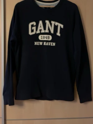 Mörkblå sweatshirt från GANT - Klassisk mörkblå sweatshirt från GANT med tryck 'GANT 1949 NEW HAVEN' i vitt på bröstet. Tröjan har rund halsringning och långa ärmar. Perfekt för en avslappnad och stilren look.