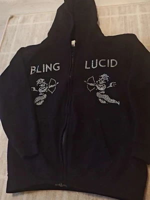 Svart hoodie med bling och text - Säljer en svart hoodie med dragkedja och huva. På framsidan står det 'BLING' och 'LUCID' i glittriga stenar, tillsammans med två cupider i samma stil. Hoodien har en avslappnad passform och är perfekt för dig som gillar statement-plagg. Storleken är 9 till 11 år!