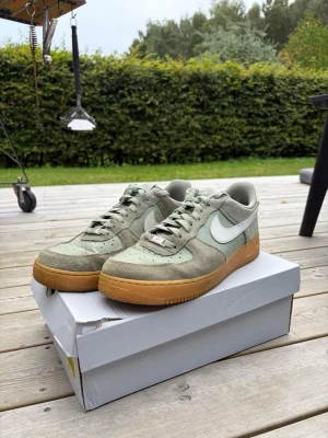 Nike Air Force 1 gröna sneakers - Snygga Nike Air Force 1 sneakers i ljusgrönt mocka och textil med klassisk vit swoosh på sidan och gummisula i brunt. Låg modell med perforerad tåbox och gröna skosnören. Perfekta för dig som vill ha en stilren och trendig look.