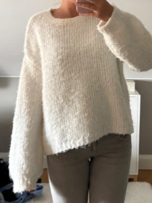 Vit fluffig stickad tröja - Mysig vit stickad tröja med oversized passform och långa ärmar. Tröjan har en rund halsringning och är gjord i ett fluffigt, mjukt material som känns riktigt skönt mot huden. Perfekt för kalla dagar när du vill vara både bekväm och snygg.