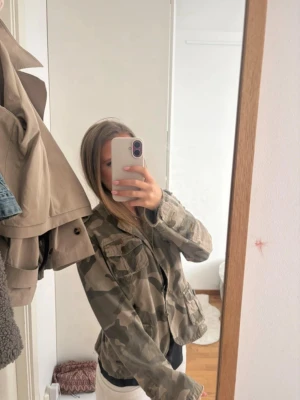 Camouflage overshirt med fickor - Säljer en snygg overshirt i camouflage-mönster med flera praktiska fickor framtill. Jackan har en avslappnad passform och stängs med knappar. Perfekt för dig som vill ha en street-inspirerad look. Materialet känns som bomull och färgerna går i grönt och beige.
