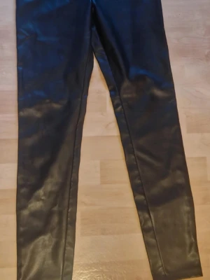 Svarta leggings i fuskskinn - Snygga svarta leggings i fuskskinn med hög midja och smal passform. Materialet ger en cool och trendig look som passar perfekt till oversized tröjor eller crop tops. Stretchigt tyg för skön komfort och enkel att styla till olika outfits.