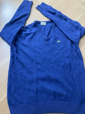 Blå V-ringad tröja från Lacoste - Mysig blå V-ringad tröja från Lacoste med den klassiska krokodilloggan på bröstet. Tröjan har långa ärmar och ribbade muddar vid ärmslut och nederkant. Perfekt för dig som gillar stilren och enkel design.