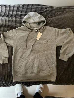 Dior hoodie - Säljer en stilren grå hoodie med huva och dragsnören. Tröjan har en broderad logga på bröstet och en stor magficka framtill. Tillverkad i mjukt sweatshirtmaterial och har ribbade muddar vid ärmar och nederkant. Perfekt för en avslappnad look.