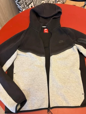 Nike Tech Fleece hoodie svart/grå - Nike Tech Fleece hoodie i svart och ljusgrått med hel dragkedja framtill. Hoodien har en huva, diskret Nike-logga på bröstet och en praktisk ficka med dragkedja på ärmen. Tillverkad i mjukt och bekvämt material som passar perfekt till streetwear.