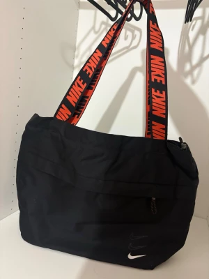 Svart väska från Nike - Snygg svart väska från Nike med breda axelband i svart och orange med Nike-logga. Väskan har dragkedja upptill, rymligt huvudfack och mindre fack framtill. Tillverkad i slitstarkt tyg, perfekt för att bära allt du behöver.