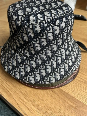 Mönstrad bucket hat i svart och beige - Snygg bucket hat med svart och beige mönster i tyg och detaljer i brunt läder runt kanten. Hatten har ett trendigt all-over print och klassisk form som passar perfekt till streetwear eller för att lyfta din outfit. ALDRIG ANVÄND