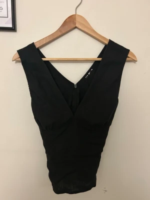 Svart v-ringad ärmlös topp - Snygg svart topp med djup v-ringning både fram och bak. Toppen är ärmlös och har en figurnära passform som framhäver siluetten. Perfekt att styla med höga jeans eller kjol för en trendig look.