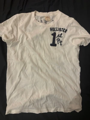 Vit Hollister t-shirt med tryck - Vit t-shirt från Hollister med mörkblå tryck på både fram- och baksidan. På bröstet står det 'Hollister 1st Pt' och på ryggen 'Surf Flyers Hollister 1st Pt'. Klassisk rund hals och korta ärmar.  tröjan är lite små klippt för design storleken är med en S/M än L 
