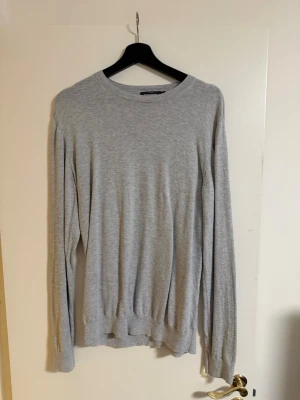 Stickad tröja Dressmann - En stilren ljusgrå tröja med rund hals och långa ärmar. Tillverkad i mjukt och tunt material, perfekt för lager på lager. Tröjan har ribbade muddar vid ärmslut och nederkant för en enkel och clean look.