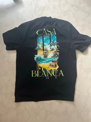 Svart Casa Blanca t-shirt med tryck - Svart t-shirt från Casa Blanca med färgglatt tryck av ett skepp, palmer och skattkista på ryggen samt mindre tryck framtill. T-shirten har klassisk passform och korta ärmar. Perfekt för dig som gillar unika motiv och streetwear-vibe.