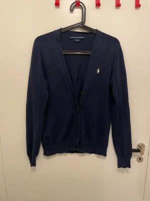 Mörkblå cardigan från Ralph Lauren - Klassisk mörkblå cardigan från Ralph Lauren med broderad logga på bröstet. Tröjan har v-ringning, knäppning framtill med svarta knappar och ribbade muddar vid ärmslut och nederkant. Perfekt att slänga över en t-shirt för en clean look.