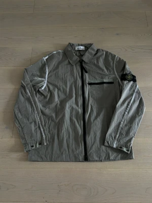 Grå overshirt med dragkedja Stone Island - Snygg grå overshirt från Stone Island med svart dragkedja framtill, bröstficka med dragkedja och ikonisk patch på vänster ärm. Overshirten har krage, knappslå vid ärmslut och detaljer längs ärmarna. Tillverkad i ett lätt, tekniskt material.