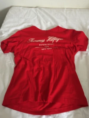 Röd Tommy Hilfiger t-shirt - Snygg röd t-shirt från Tommy Hilfiger med vit logga och texten 'Established 1985 New York' framtill. Klassisk passform och rund halsringning. Perfekt till jeans eller shorts för en avslappnad look.
