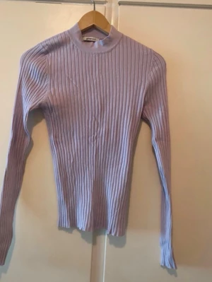 Ljuslila ribbstickad tröja - Säljer en ljuslila ribbstickad tröja med lång ärm och hög hals. Tröjan har en tight passform och är gjord i ett mjukt, stretchigt material som sitter snyggt på kroppen. 