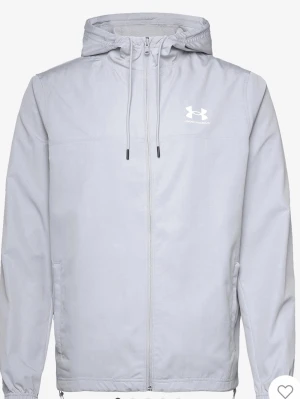 Ljusgrå vindjacka från Under Armour - Snygg ljusgrå vindjacka från Under Armour med huva och dragsko. Jackan har hel dragkedja framtill, två sidofickor och elastiska muddar vid ärmsluten. Tillverkad i lätt och vindtätt material, perfekt för blåsiga dagar.