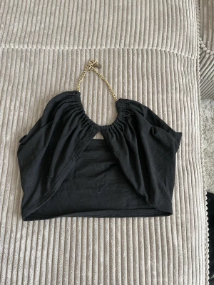 Svart axelbandslös topp med kedja - Trendig svart axelbandslös topp med guldfärgad kedja som halterneck. Toppen har en öppen design framtill och är croppad, perfekt för att sticka ut. Materialet känns mjukt och följsamt, och toppen har en cool, modern vibe.
