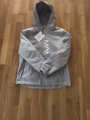 Ljusgrå vindjacka med huva - Snygg ljusgrå vindjacka från Moncler med stor huva och dragkedja framtill. Jackan har två sidofickor med dragkedja och justerbar nederkant. Materialet är tunt och vindtätt, perfekt för blåsiga dagar. Stor logga framtill ger en modern look.