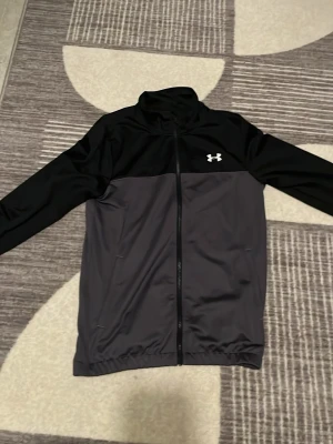 Svart och grå träningsjacka Under Armour helt ny köptes igår  - Snygg träningsjacka från Under Armour i svart och mörkgrått. Jackan har hel dragkedja, ståkrage och två sidofickor. Tillverkad i ett lätt och stretchigt funktionsmaterial som passar perfekt till träning eller uppvärmning.