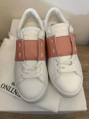 Valentino vita sneakers med rosa detalj - Snygga vita sneakers från Valentino med bred rosa läderdetalj över snörningen. Klassisk låg modell med rund tå och vita skosnören. Perfekta för dig som vill ha en stilren men ändå unik look.