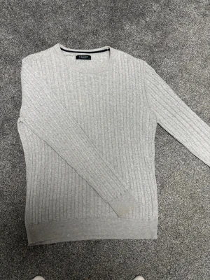 Ljusgrå kabelstickad tröja - En stilren ljusgrå kabelstickad tröja med rund halsringning och långa ärmar. Tröjan har ribbade muddar vid ärmslut och nederkant, vilket ger en snygg och klassisk look. Perfekt att matcha med jeans eller kjol för en enkel men trendig outfit.