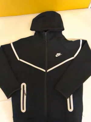 Svart Nike hoodie med reflexdetaljer - Snygg svart hoodie från Nike med dragkedja framtill och vita reflexdetaljer längs bröstet och vid fickorna. Passar som xs. Klassisk huva och två fickor med dragkedja. Nike-logga på bröstet. Perfekt för dig som gillar sportig stil och vill synas i mörkret.