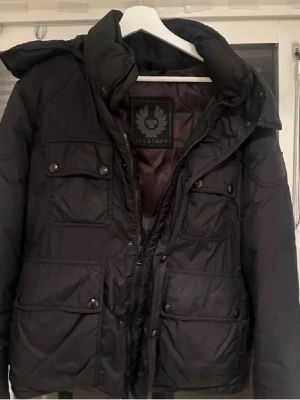 Svart pufferjacka från Belstaff - Säljer en svart pufferjacka från Belstaff med fyra framfickor med knappar och hög krage. Jackan har en klassisk quiltad design och dragkedja med knappslå. Perfekt för kalla dagar och riktigt snygg till streetwear.