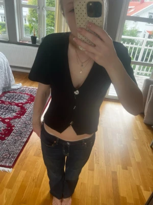 Svart v-ringad topp med knappar - Snygg svart topp med v-ringning och korta ärmar. Toppen har dekorativa knappar framtill och en figurnära passform som ger en stilren look. Perfekt att matcha med både jeans och kjol för en trendig outfit.