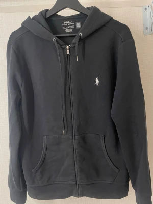 Svart hoodie från Polo Ralph Lauren - Klassisk svart hoodie från Polo Ralph Lauren med dragkedja, huva med dragsko och fickor framtill. Ikoniska vita logon broderad på bröstet. Tillverkad i mjukt bomullsmaterial som är skönt att bära. Perfekt för en avslappnad och stilren look.