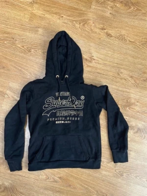 Svart Superdry hoodie med tryck - Svart hoodie från Superdry med stort vitt tryck på bröstet och japanska tecken. Klassisk känguruficka framtill, dragsko i huvan och ribbade muddar. Perfekt för en avslappnad och cool stil.