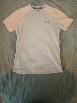 Vit t-shirt från Under Armour - Säljer en vit t-shirt från Under Armour med klassisk passform och diskret logga på bröstet. T-shirten har korta ärmar och rund halsringning. Perfekt för dig som gillar enkel och sportig stil.