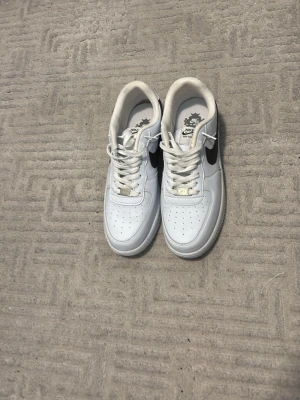 Vita Nike Air Force 1 sneakers - Klassiska vita Nike Air Force 1 sneakers med svart swoosh på sidan. Skorna har lågt skaft, perforerad tåbox och vit sula med cirkulärt mönster undertill. Tillverkade i läder med vita skosnören och diskreta detaljer. Perfekta för en clean och stilren look.