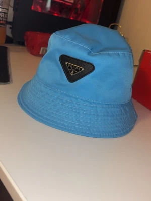 Blå bucket hat  - Snygg blå bucket hat med triangulär logga framtill i svart och silver. Hatten är tillverkad i ett glansigt material som ger en modern vibe och passar perfekt till streetwear eller sommarens outfits. QR kod som funkar!