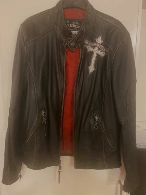 Herr Svart skinnjacka från Affliction storlek M - Riktigt snygg svart skinnjacka från Affliction med coola broderade detaljer. Framsidan har ett korsmärke på bröstet och baksidan har ett stort broderi med kors, dödskalle och vingar. Röd innerfoder och dragkedjor på fickorna. 