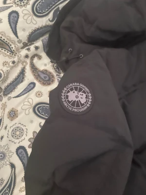 Svart pufferjacka från Canada Goose - Säljer en svart pufferjacka från Canada Goose med klassisk patch på ärmen. Jackan har huva med dragsko och breda, vadderade paneler för extra värme. Perfekt för kalla vinterdagar och riktigt snygg streetstil.