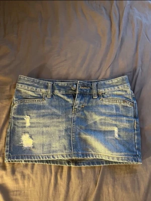 Lågmidjad Blå jeanskjol med slitningar - Snygg blå jeanskjol i kort modell med slitna detaljer framtill. Kjolen har klassiska fickor både fram och bak samt hällor för skärp. Perfekt för en avslappnad och trendig look. Köpt på vinted men var tyvär för liten för mig, gamla ägarens bilder. Midjemåttet är 39cm och längden är 26cm