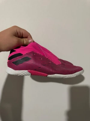 Rosa fotbollsskor med svarta detaljer - Säljer ett par riktigt snygga fotbollsskor i stark rosa färg med svarta ränder och detaljer. Skorna har en låg profil och vit sula som passar perfekt för inomhusspel. Tillverkade i syntetmaterial för bra passform och känsla på planen.