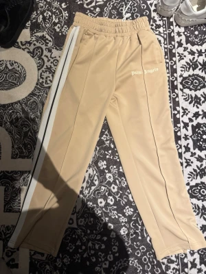 Beige mjukisbyxor Palm Angels - Snygga beige mjukisbyxor från Palm Angels med vita sidoränder och logga på vänster sida. Byxorna har resår i midjan och raka ben. Perfekta för en chill och trendig look.