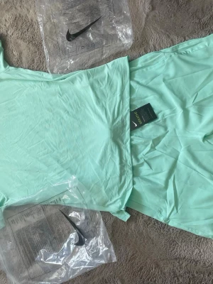 Mintgrön sporttröja från Nike - Snygg mintgrön sporttröja från Nike med klassisk logga på bröstet. Tillverkad i ett lätt och snabbtorkande material som passar perfekt till träning. Tröjan har rund hals och korta ärmar för maximal rörelsefrihet.