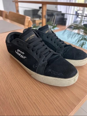 Yves Saint Laurent shoes - Säljer ett par svarta sneakers från Saint Laurent med vit logga på sidan och guldfärgad text på plösen. Skorna har svart tyg och läderdetaljer, samt vita sulor. Klassisk låg modell med svarta skosnören och stilren design.