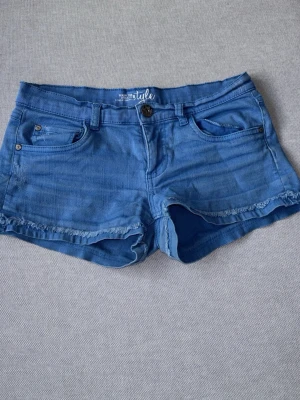 Blå jeansshorts  - Säljer ett par blå jeansshorts med rå kant nertill och klassisk femficksdesign. Shortsen har normal passform och bälteshällor i midjan. Perfekta för varma dagar och enkla att matcha med olika toppar. De är i bra skick men lite urtvättade men det är inget man tänker på!