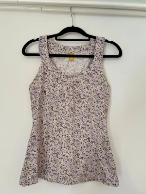 Blommig ärmlös linnetopp i lila - Supersöt ärmlös linnetopp med blommigt mönster i lila, grönt och vitt. Toppen har rund halsringning och är lätt utsvängd nertill, perfekt för varma dagar. Tillverkad i mjuk bomull som känns skön mot huden.