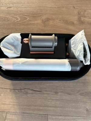 Dyson hårstylingverktyg med tillbehör - Säljer ett hårstylingverktyg från Dyson med flera tillbehör. Setet innehåller olika munstycken och kommer i en mörkblå förvaringsväska. Materialet är mestadels metall och plast, perfekt för dig som vill fixa håret med smarta verktyg.