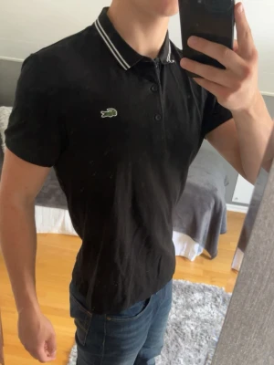 Svart pikétröja med vit krage - Säljer min svarta Lacoste piké.  Den har inga fläckar eller skador, kontakta mig gärna vid funderingar 