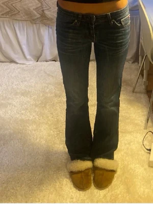 Mörkblå bootcut jeans - Säljer ett par mörkblå bootcut jeans med klassisk femficksdesign och låg midja. Jeansen har en lätt tvättad look och är tillverkade i ett stretchigt denimtyg som sitter snyggt över höfterna och låren innan de släpper ut vid bensluten.