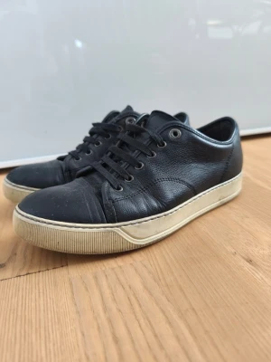 Lanvin skor - Snygga svarta sneakers i läder/skinn med vita sulor och svarta skosnören. Passar perfekt till jeans eller shorts för en clean och stilren look. Skick 6/10. Skada på sista bilden.                     En del slitage och creasade men fullt fungerande och inga hål osv...                        Med lite ny skofärg och tvättning kan det bli riktigt fina igen. Mitt pris 750kr.                       Pris kan diskuteras!